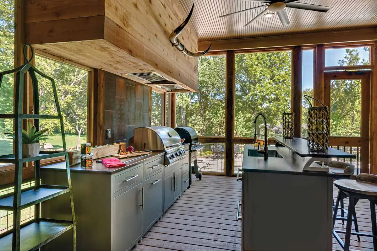 Outdoor kitchen with stainless steel grills, counter space, sink, and decorative lanterns. Shelving holds plants and spices. Large windows offer a view of lush greenery.
