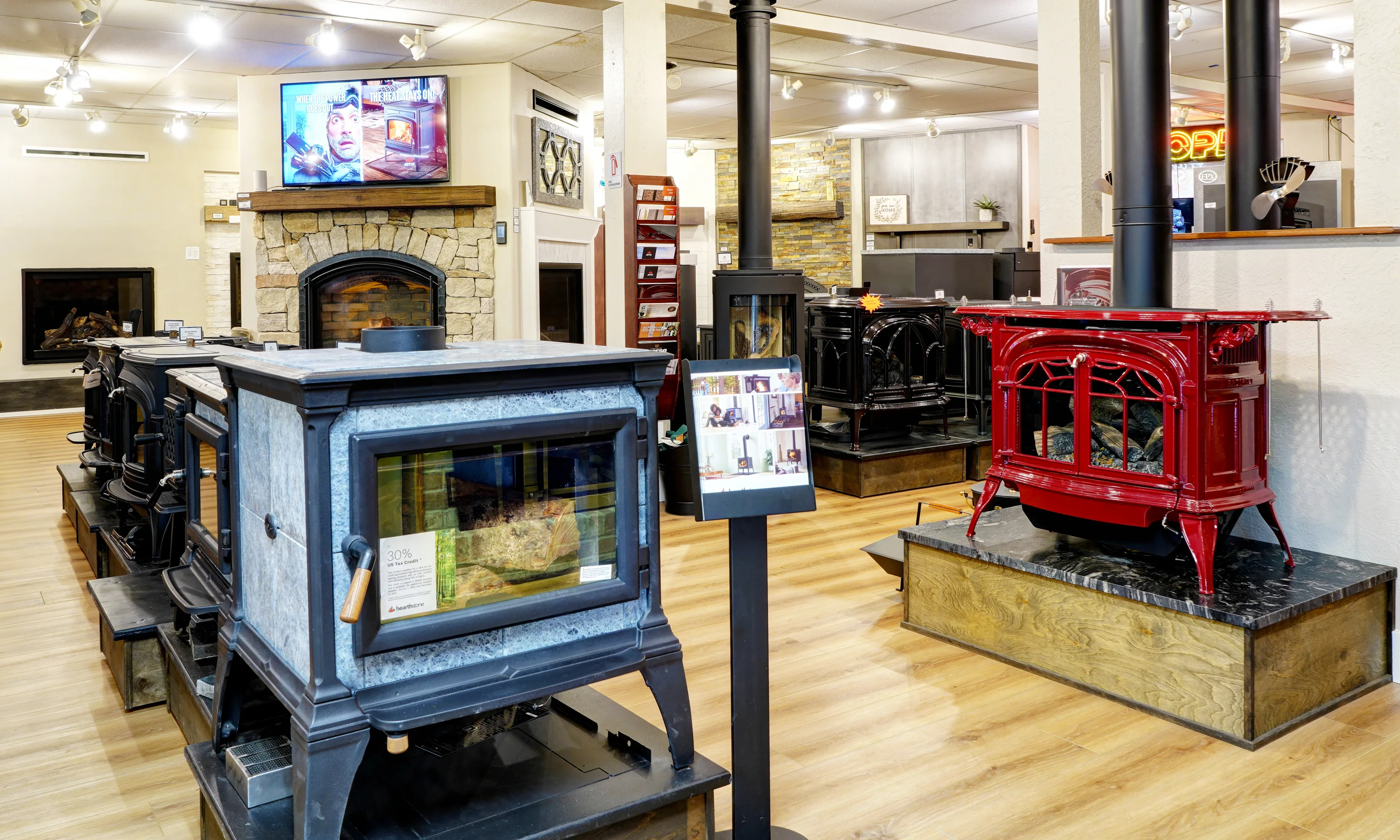 Showroom displaying a variety of freestanding wood-burning stoves and a stone fireplace, including a prominent red cast-iron stove on a raised platform.