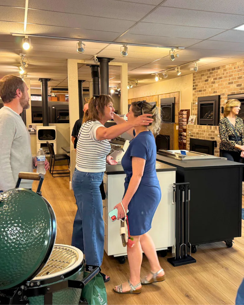 Two women greet each other with a hug in a cozy store, surrounded by various home appliances and decor. Others engage nearby under warm lighting.