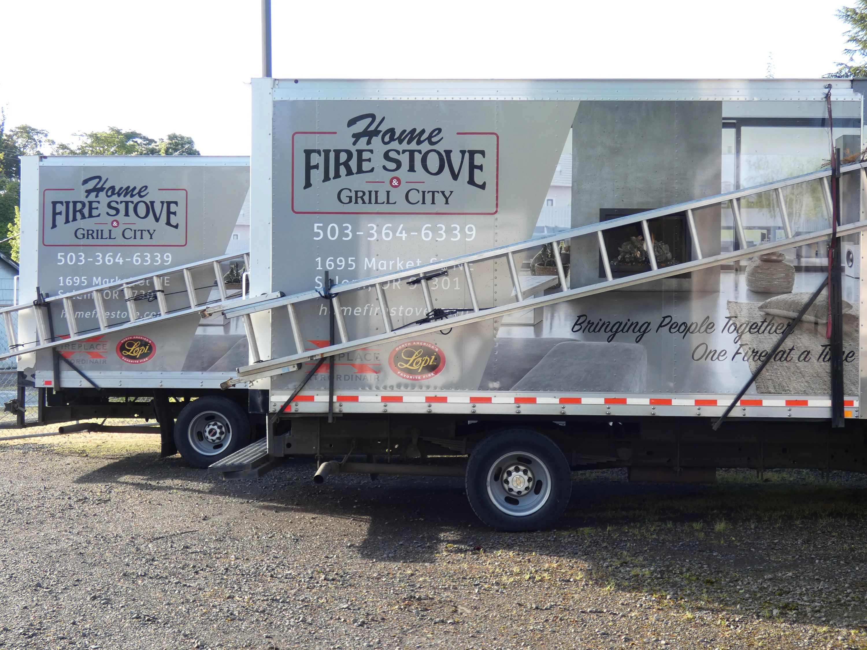 Two trucks with Home Fire Stove Grill City branding, displaying contact information and an image of a cozy living room. Ladders are mounted on the truck sides.