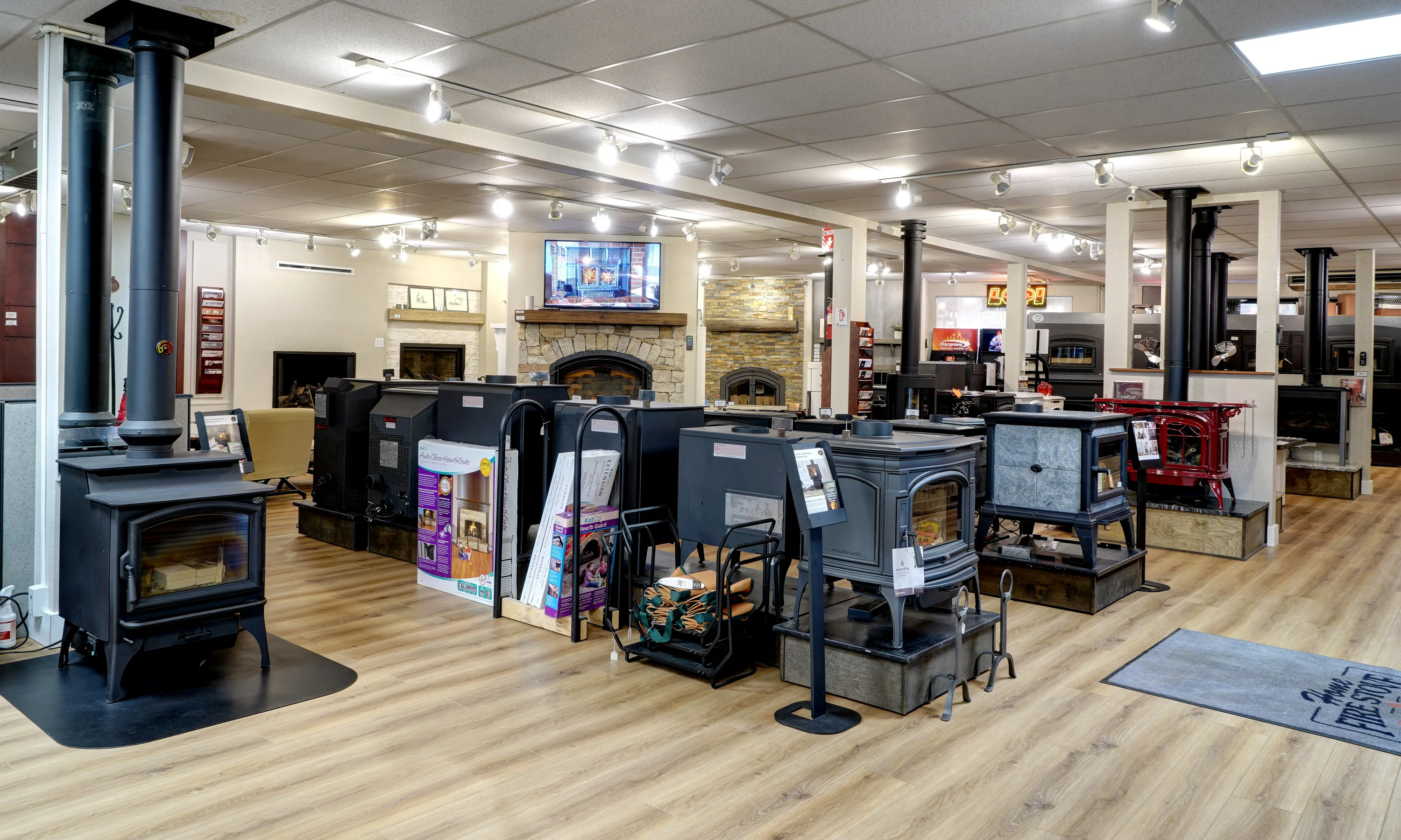 A showroom with various models of stoves and fireplaces displayed. The room has wooden flooring, overhead lighting, and a television mounted above a stone fireplace in the background.