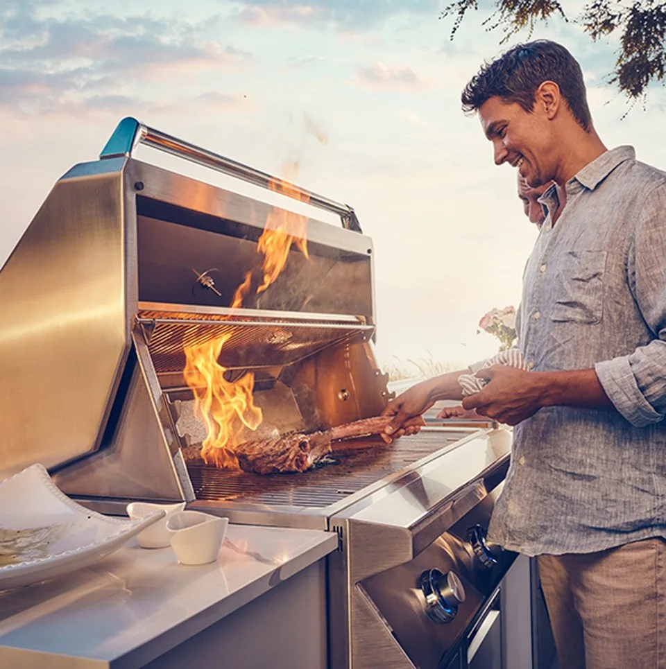 Man using tongs to grill a large steak on a stainless steel gas grill with open hood and visible flames, smiling outdoors at sunset