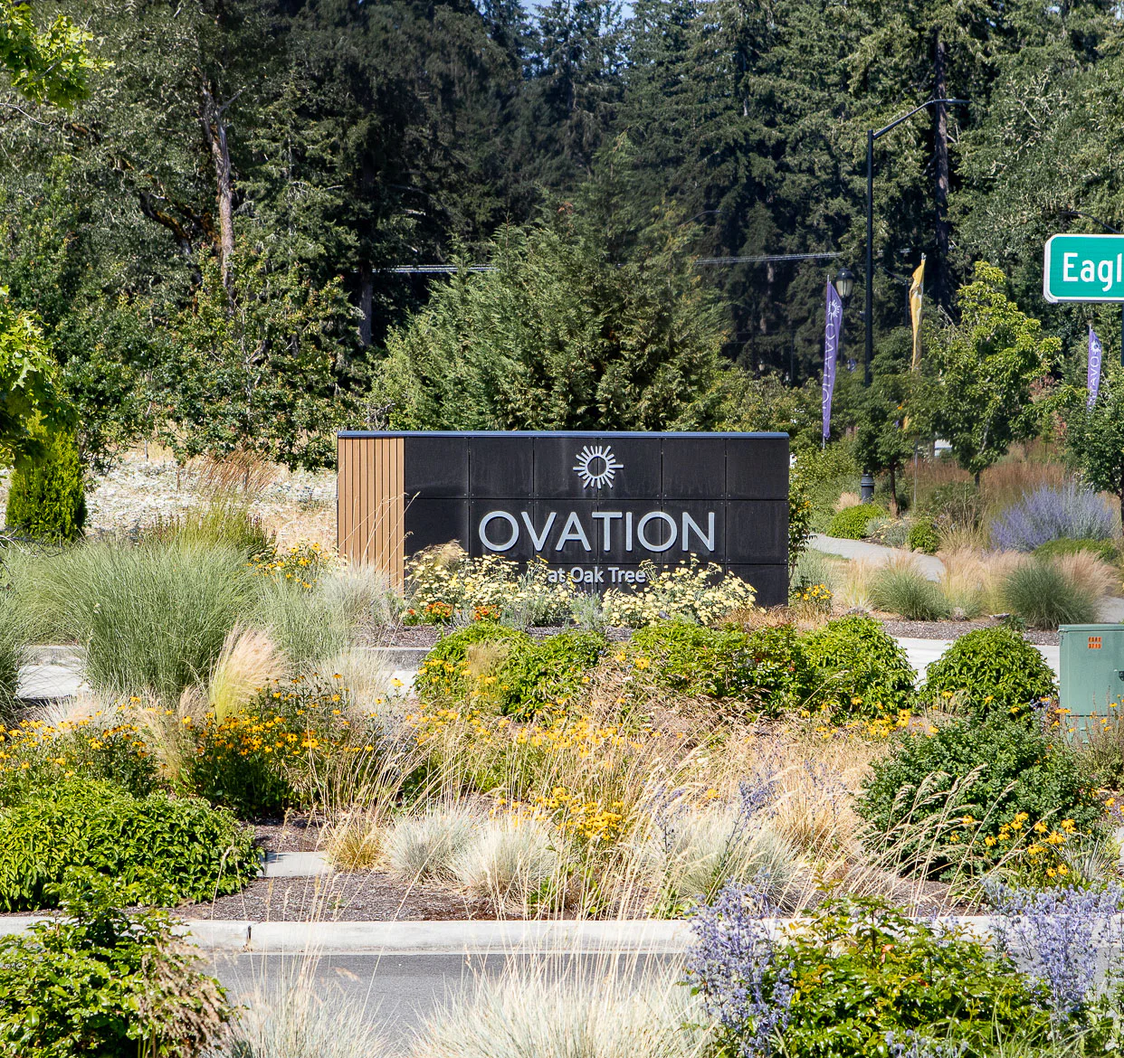 A sign reads Ovation at Oak Tree, surrounded by landscaped greenery and colorful flowers, with tall trees and a street sign for Eagle nearby.