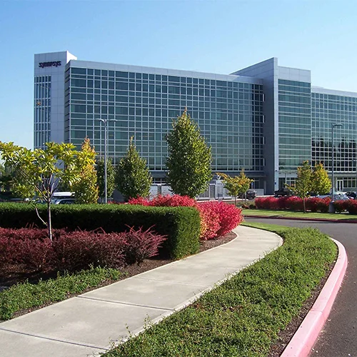 Modern glass office building with multiple stories, surrounded by a landscaped garden with green shrubs, red bushes, and young trees. A curved sidewalk leads to the building entrance.