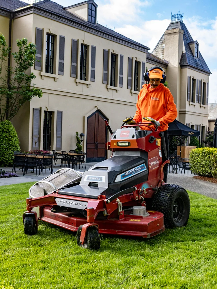 A person in an orange hoodie and cap operates a red Toro Grandstand lawn mower on a neatly manicured lawn in front of a large, light-colored building with many windows and outdoor tables.
