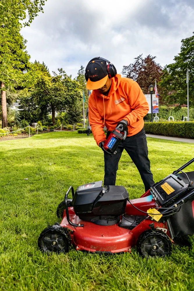 A person wearing an orange hoodie, black pants, ear protection, and a cap is refueling a red lawnmower on a grassy lawn, surrounded by trees and flags in the background.
