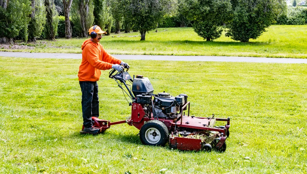 A person wearing orange safety gear and earmuffs operates a red riding lawn mower in a park with green grass and trees in the background.