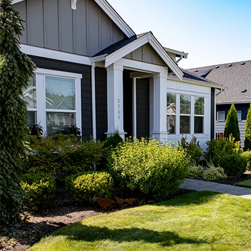 Gray house with white trim featuring a covered porch and surrounded by a well-maintained garden with various shrubs and small trees. The house number 3739 is visible on a porch column.