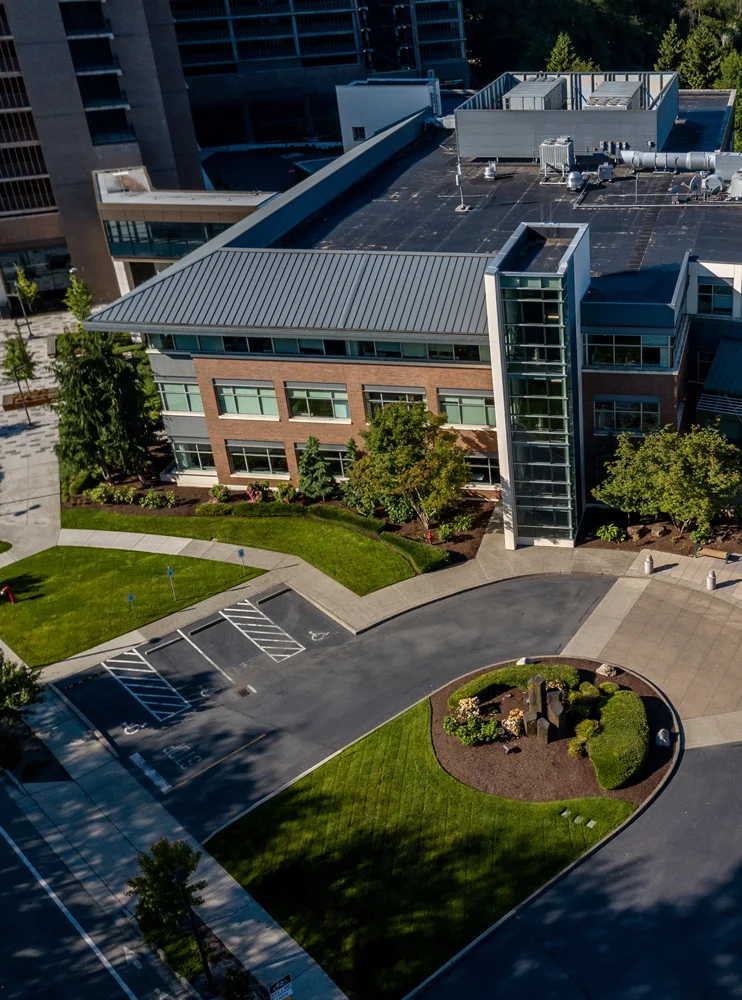 Aerial view of a contemporary office building with large windows and a sloped metal roof. The building is surrounded by green lawns, trees, and a roundabout with landscaping features.