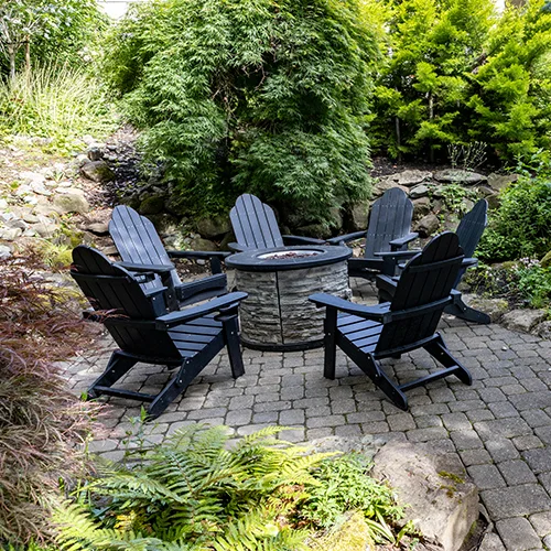 A patio with six black Adirondack chairs arranged in a circle around a stone fire pit, surrounded by lush green plants and large trees.