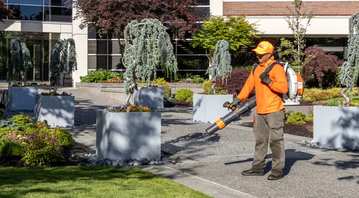 A landscaper wearing an orange shirt and hat operates a leaf blower in a well-maintained garden with large square planters, greenery, and a modern building in the background on a sunny day.