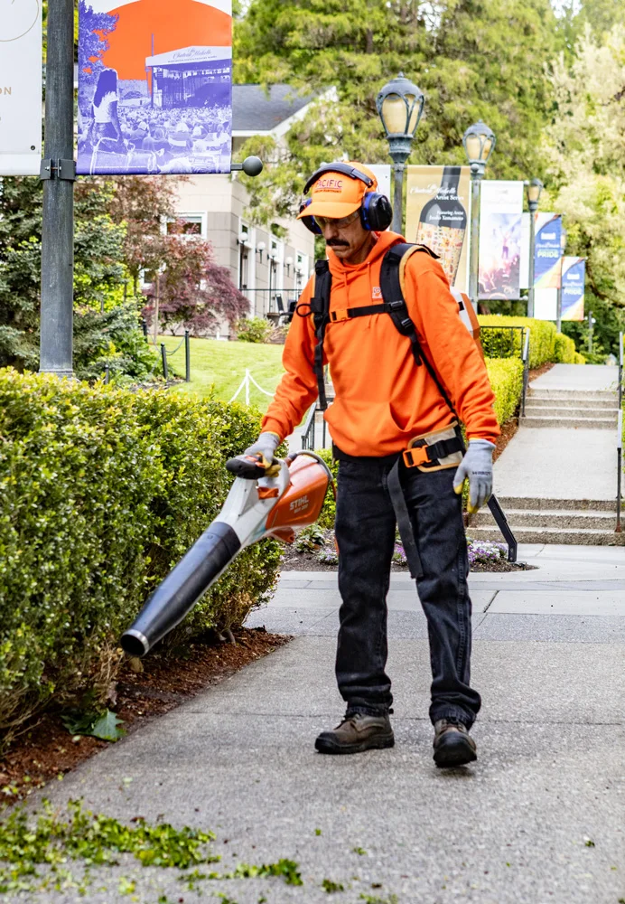 Worker in an orange jacket and protective gear uses a leaf blower to clear a sidewalk surrounded by greenery. Banners hang from poles along the path, and a building is visible in the background.