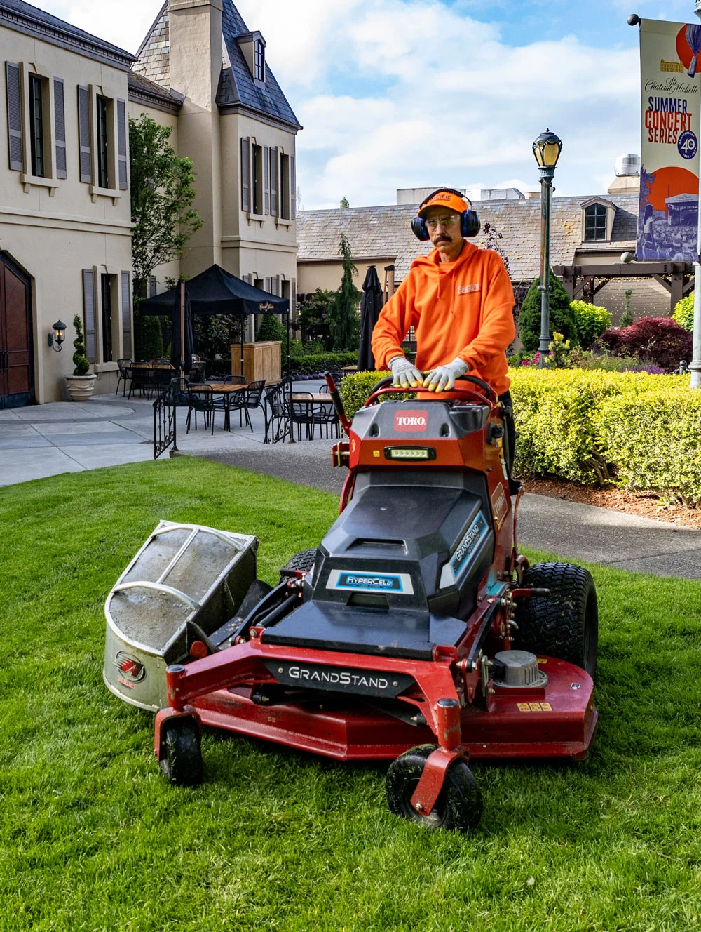 A person wearing an orange hoodie and ear protection mows the lawn with a Toro GrandStand lawnmower in front of a building with beige walls and several windows. A