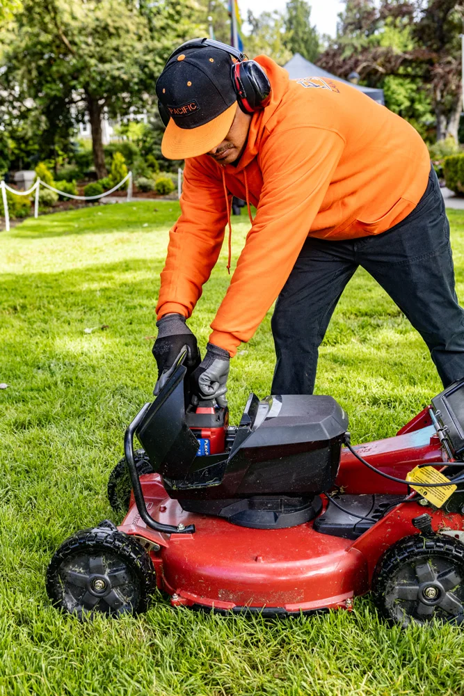 A person in an orange hoodie and black hat, wearing ear protection, is standing on a grassy lawn and replacing the battery in a red lawnmower. Trees and bushes are in the background.