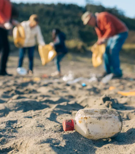 A plastic bottle lies on the sand in the foreground, while a group of people in the background pick up trash on a beach.