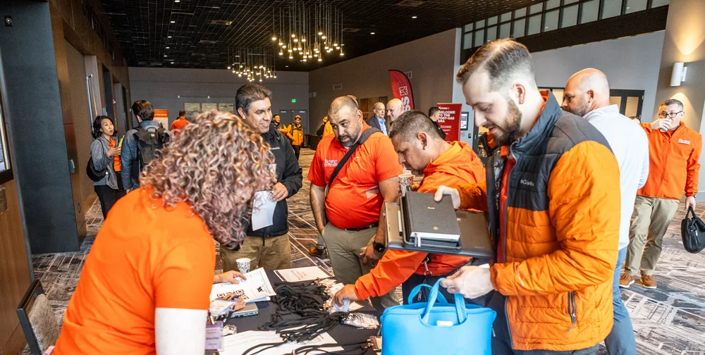 People gather around a table with brochures and lanyards during a networking event in a well-lit conference room. Some participants wear orange shirts and jackets.
