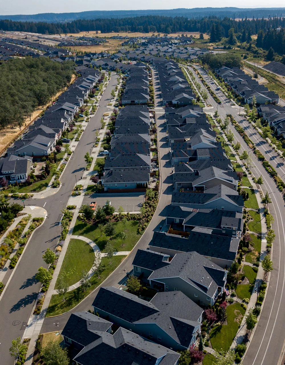 Aerial view of a suburban neighborhood with rows of houses, tree-lined streets, and green spaces. The area is surrounded by forests and fields, extending towards the horizon.