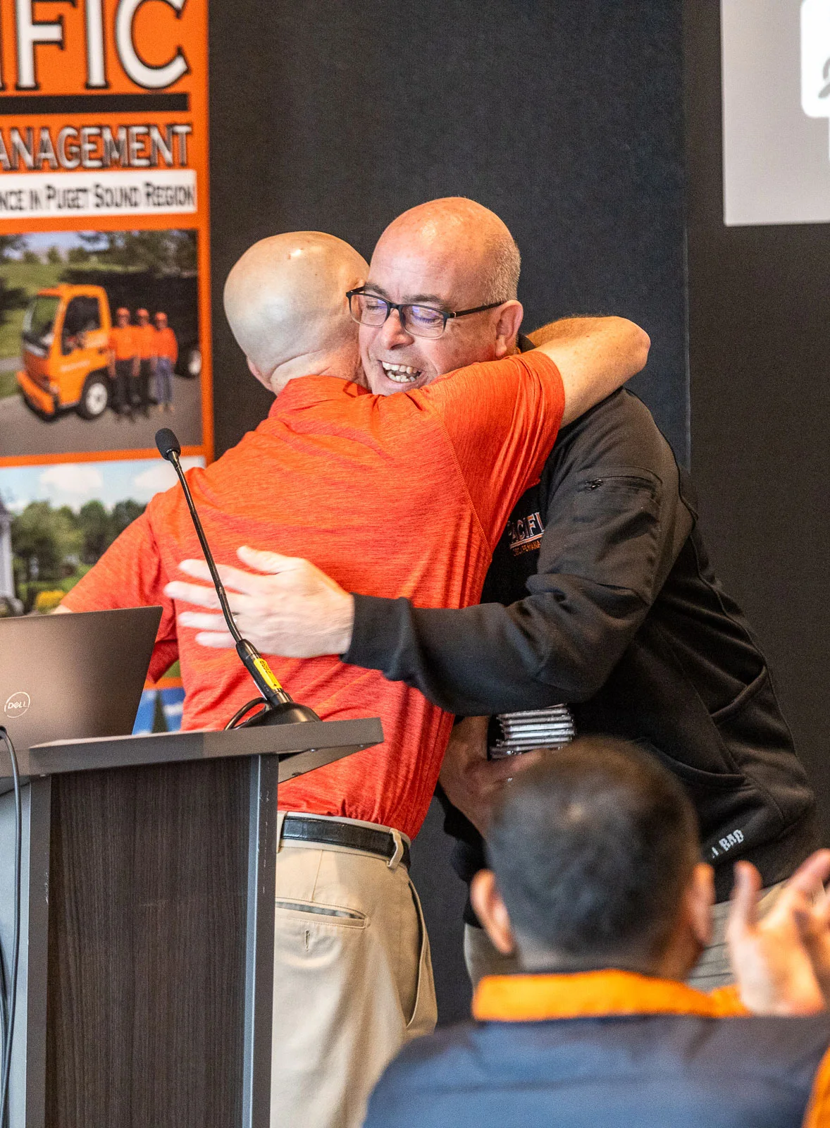 Two men hug during a presentation, one in an orange shirt at a podium, the other in a black jacket. A banner behind them reads
