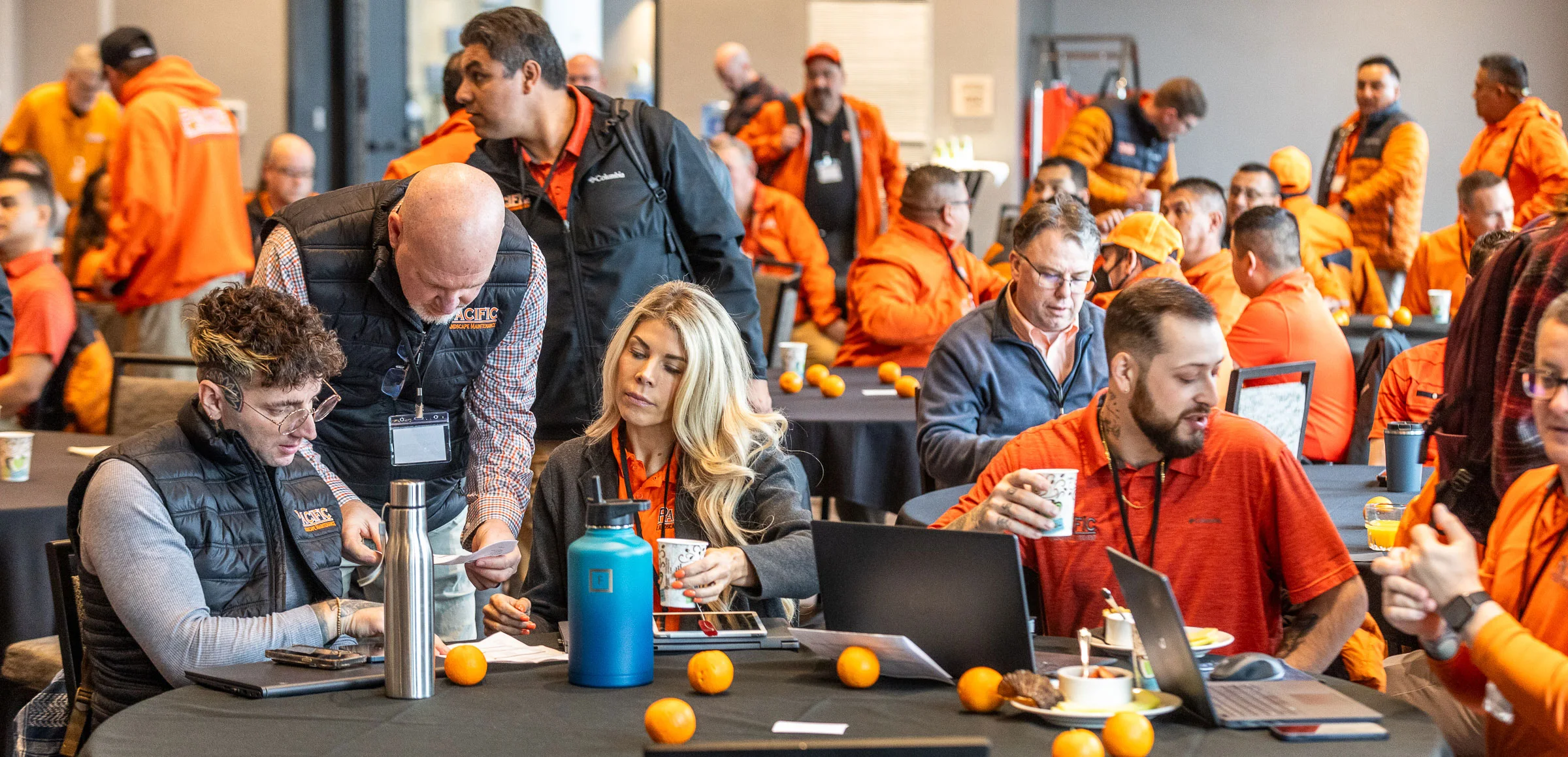 A group of people in orange and black attire are attending a meeting. Some are seated at tables with laptops and cups, engaging in discussions, while others are standing or walking around.