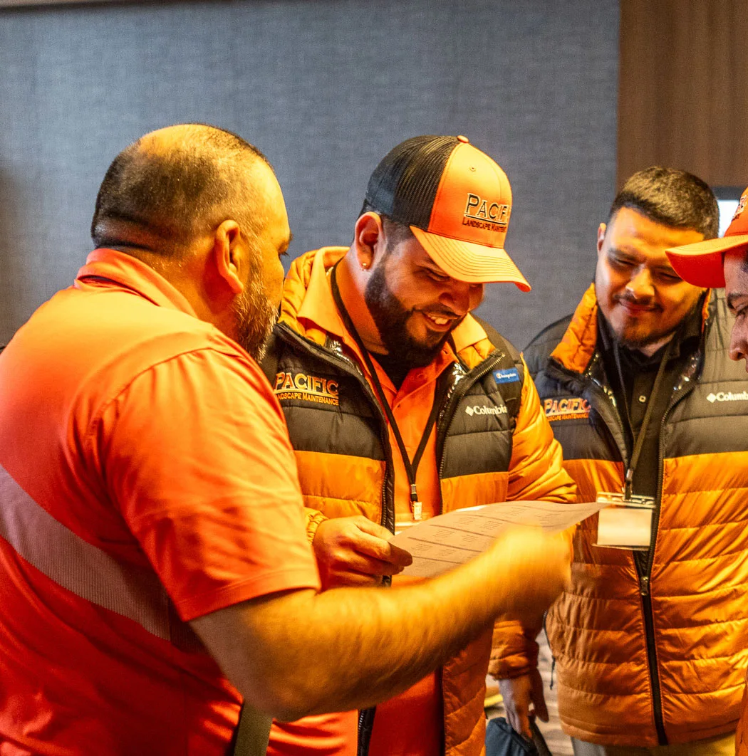 Four men wearing orange shirts and jackets with company logos are gathered together, smiling and looking at a document. They appear to be discussing something work-related.