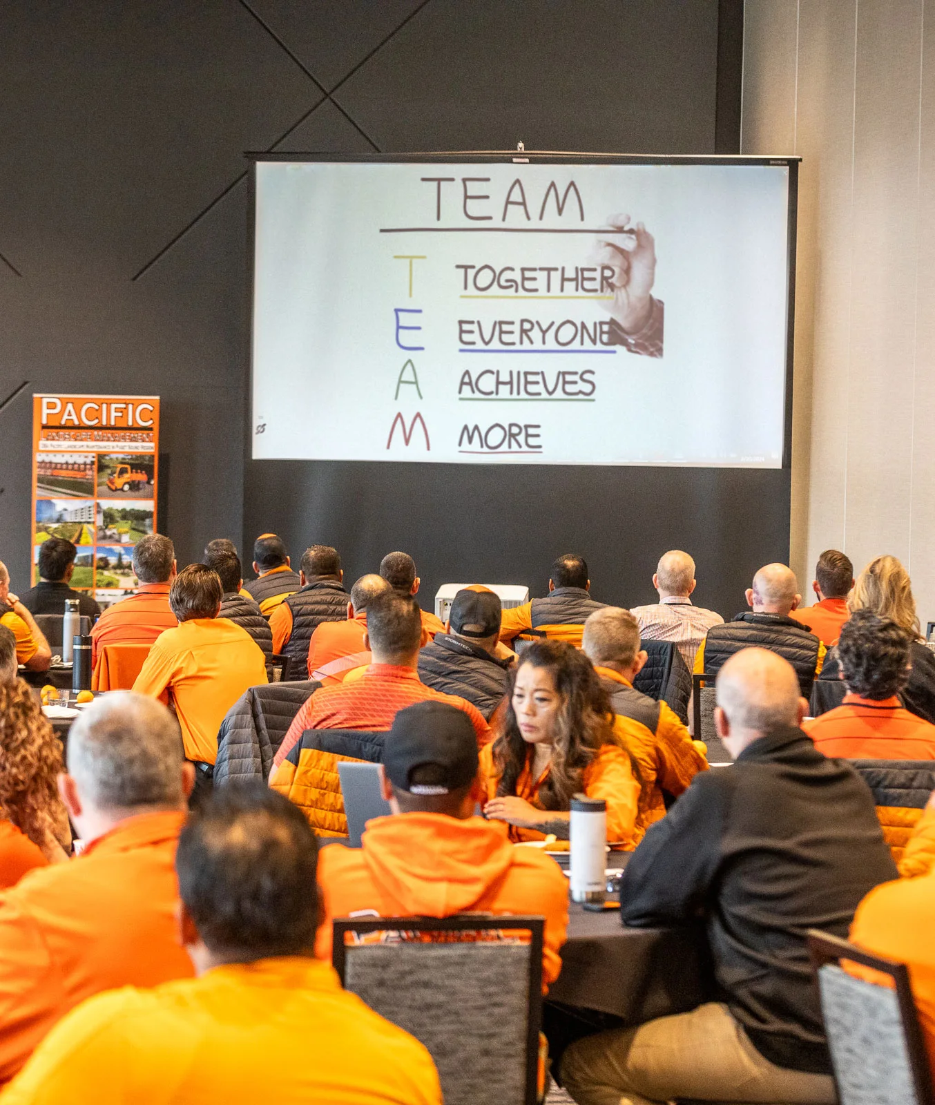 A group of people, mostly in orange jackets, sitting in a conference room looking at a screen displaying