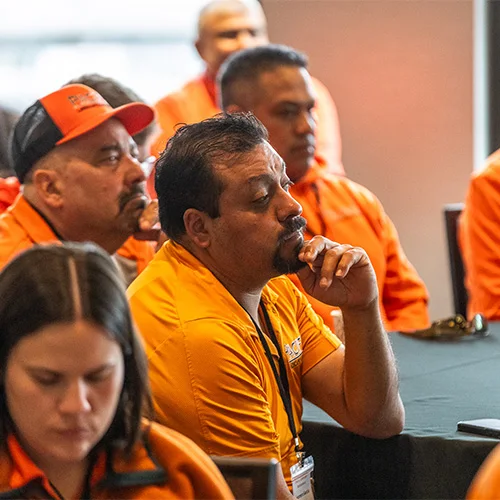 Group of people wearing orange shirts and lanyards sitting at a table during a meeting or conference. Man in the foreground rests his chin on his hand, appearing thoughtful.
