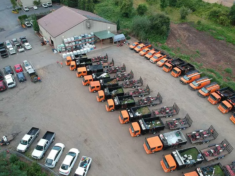An aerial view of a commercial parking lot with two rows of orange trucks with trailers parked in an open area and several white and black vehicles parked next to a building.