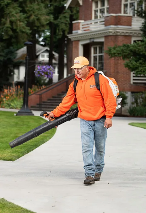 A man in an orange hoodie, jeans, and a beige cap uses a leaf blower on a sidewalk in front of a brick building with large windows and greenery around.