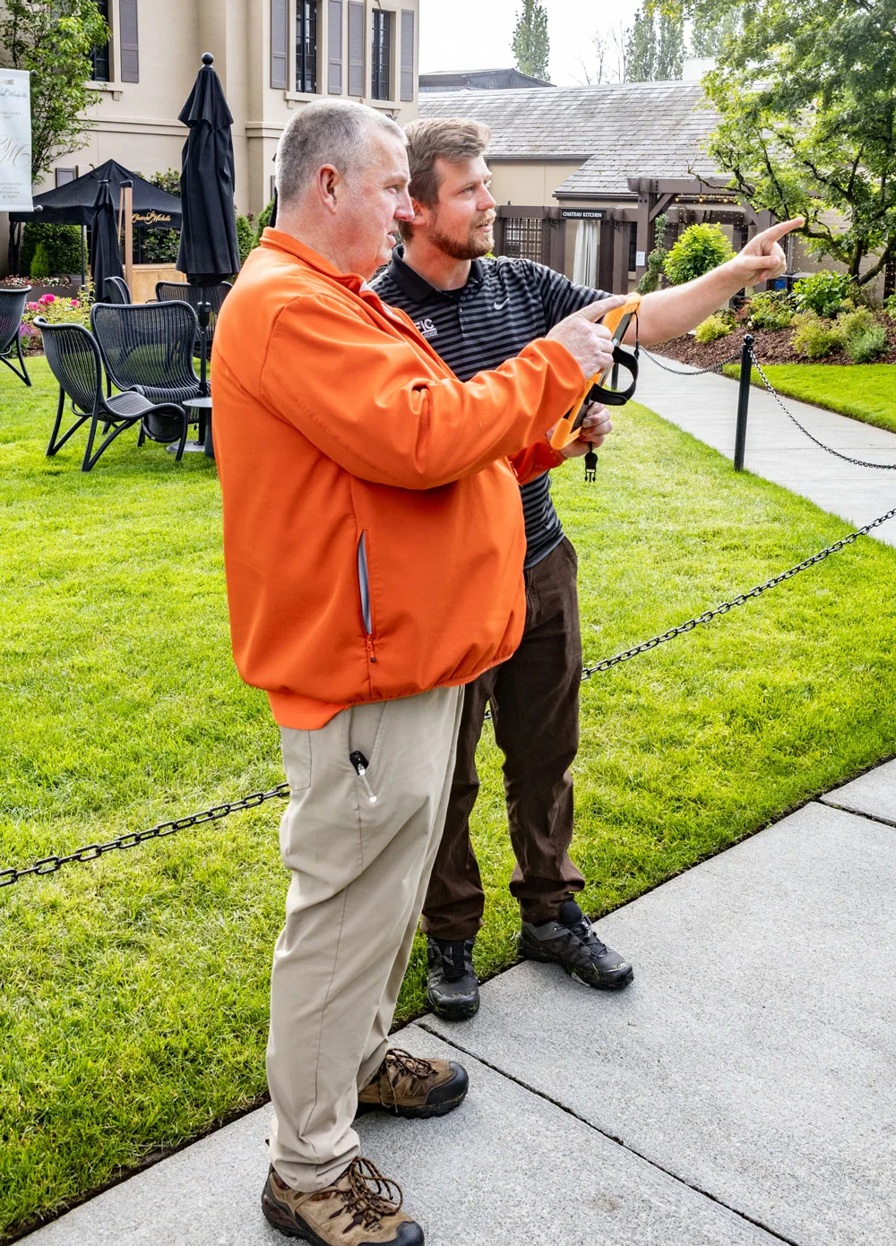 Two men, one in an orange jacket and another in a striped shirt, stand on a manicured lawn and point while holding documents. They appear to be discussing something outdoors.