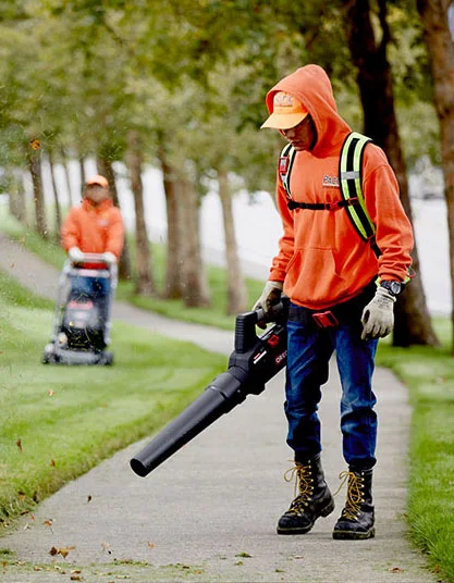 Two workers in orange hoodies and reflective vests maintain a park. One uses a leaf blower on a path, while the other operates a lawn mower in the background.