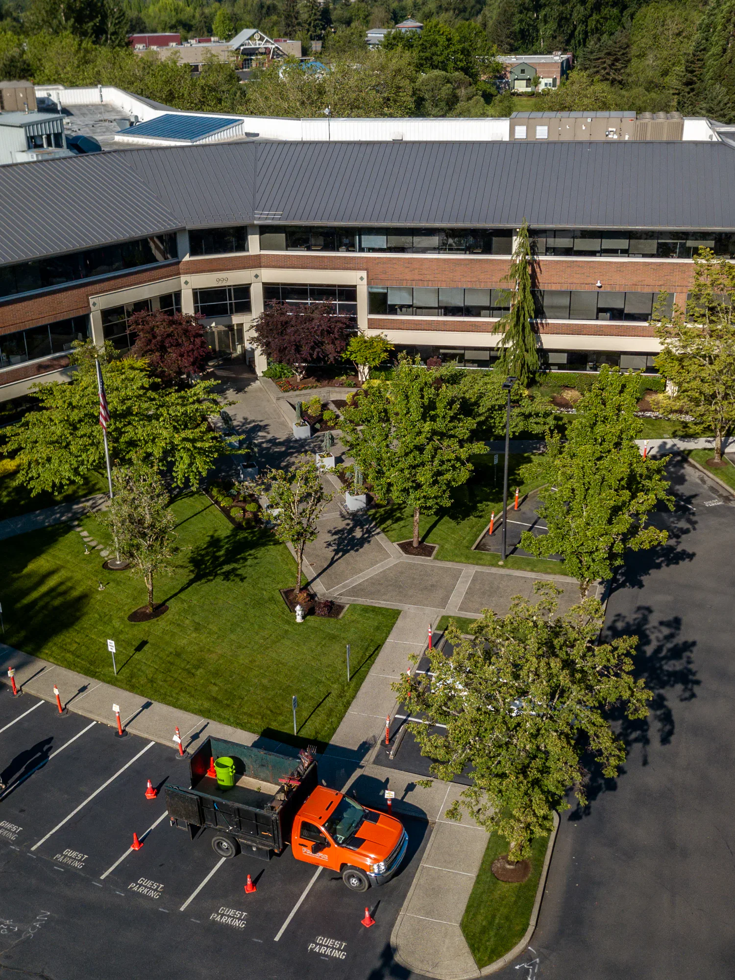 Aerial view of a two-story office building with a grey roof, surrounded by trees and greenery. An orange utility truck is parked near guest parking spaces marked by traffic cones.