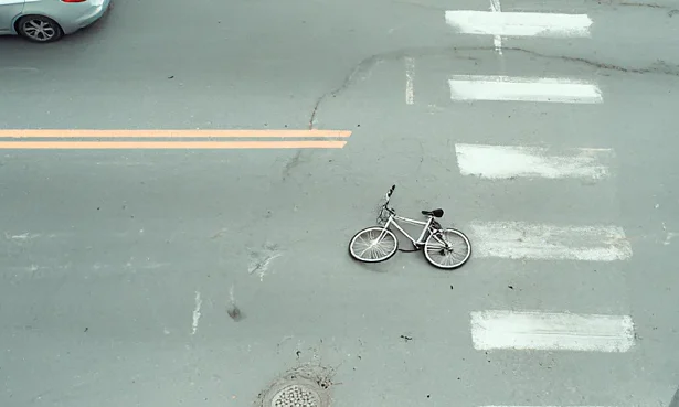 Aerial view of a quiet street intersection with a silver bicycle lying on its side in the middle of a crosswalk. A car is partially visible on the left side of the image.