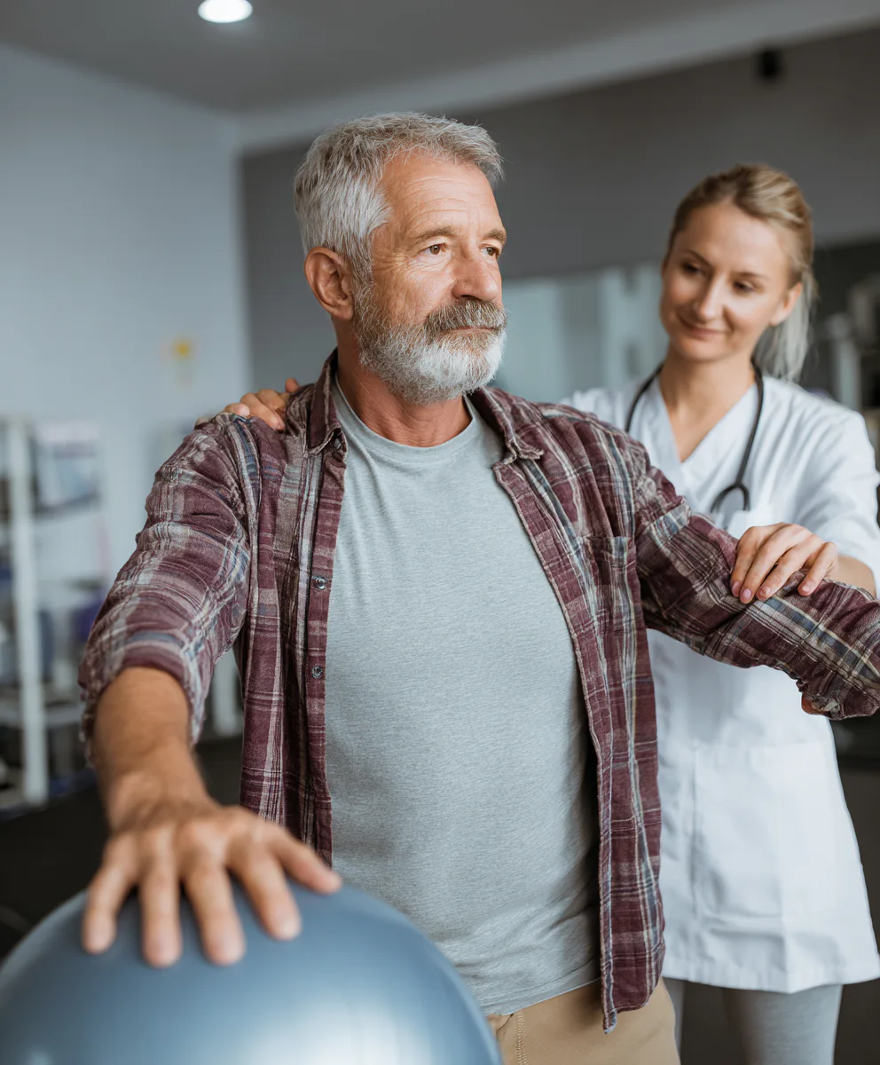 Elderly man doing rehabilitation exercises with a stability ball, assisted by a healthcare professional in a medical setting. The man is wearing a plaid shirt and has gray hair.
