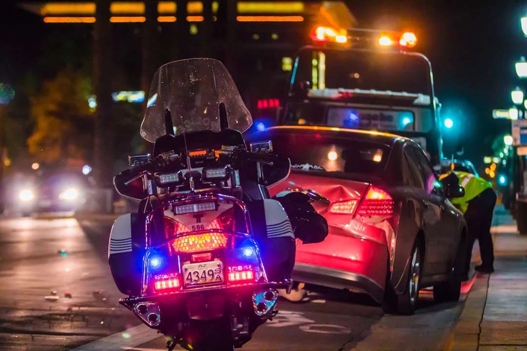 A police motorcycle with flashing lights is parked on a nighttime street behind a damaged car and tow truck. A person in a reflective vest inspects the scene.