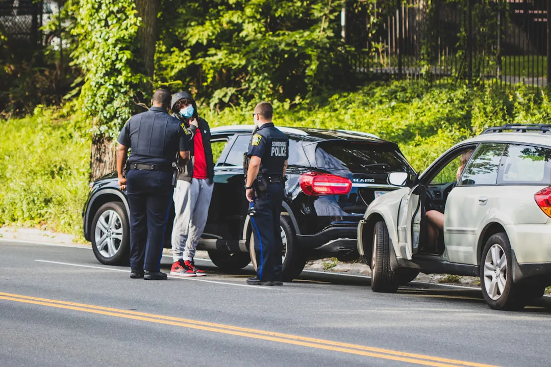 Police officers standing by a black SUV talking to a masked person in a red shirt on a roadside. Nearby, someone is seated in a parked white car with an open door.