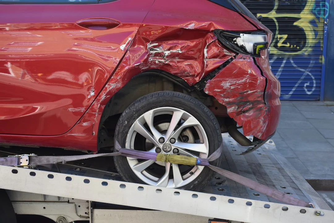 A red car with significant damage to the rear bumper and side panel is secured on a tow truck with straps. The car's wheel and scratched paint are visible.