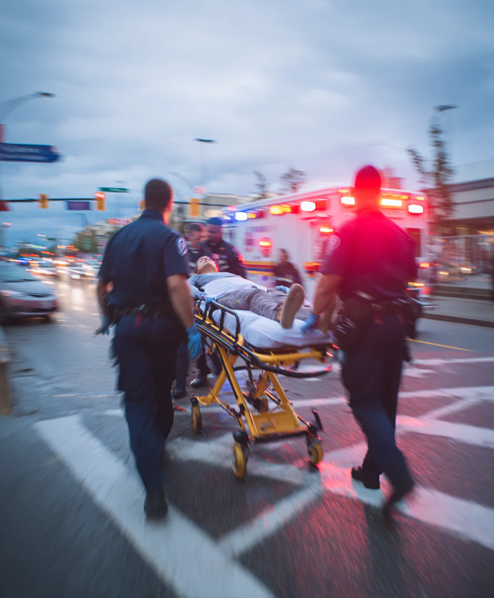 Emergency responders push a stretcher with a person towards an ambulance with flashing lights at dusk, on a busy street with cars and traffic signals visible.