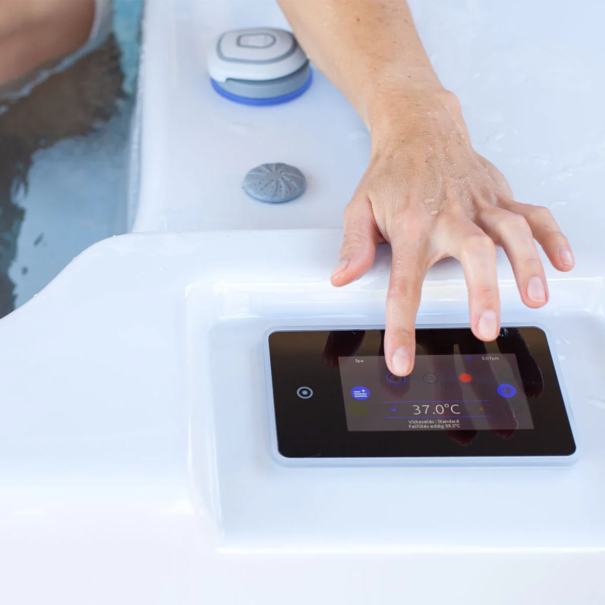 Close-up of a hand touching a hot tub touchscreen control displaying 37.0 C on the spa panel, with wet tub edge and water buttons nearby.