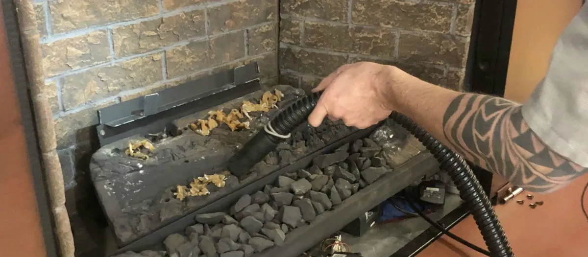 A person with a tattooed arm using a vacuum cleaner attachment to clean a fireplace with black rocks and some yellow debris inside. The fireplace has a brick interior.