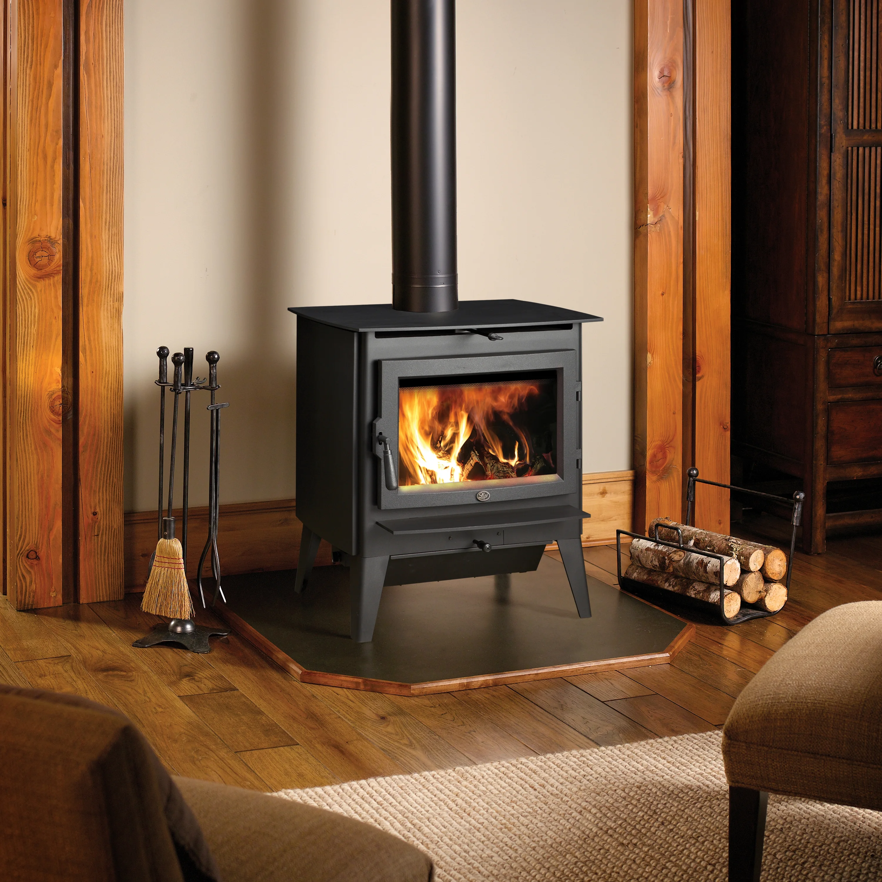 A cozy room with a lit black wood-burning stove, surrounded by wooden decor. To the left are fireplace tools and to the right is a log holder. A brown chair is in the foreground.