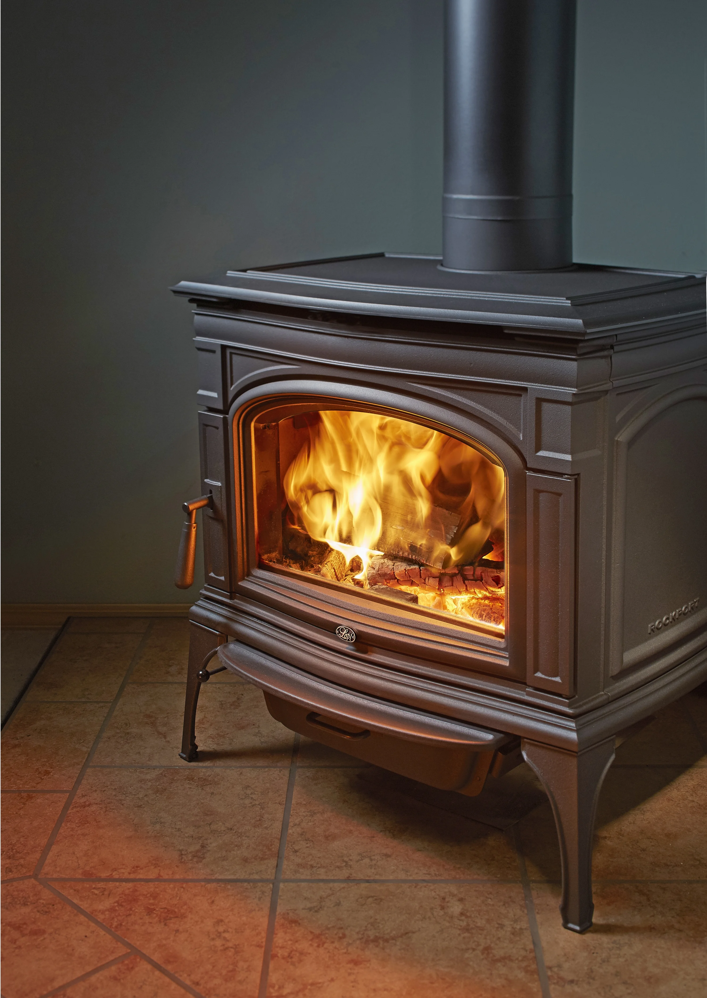 A black cast iron wood stove with a glass front, showing a bright, roaring fire inside. The stove is placed on a tiled floor, illuminating the surrounding area.