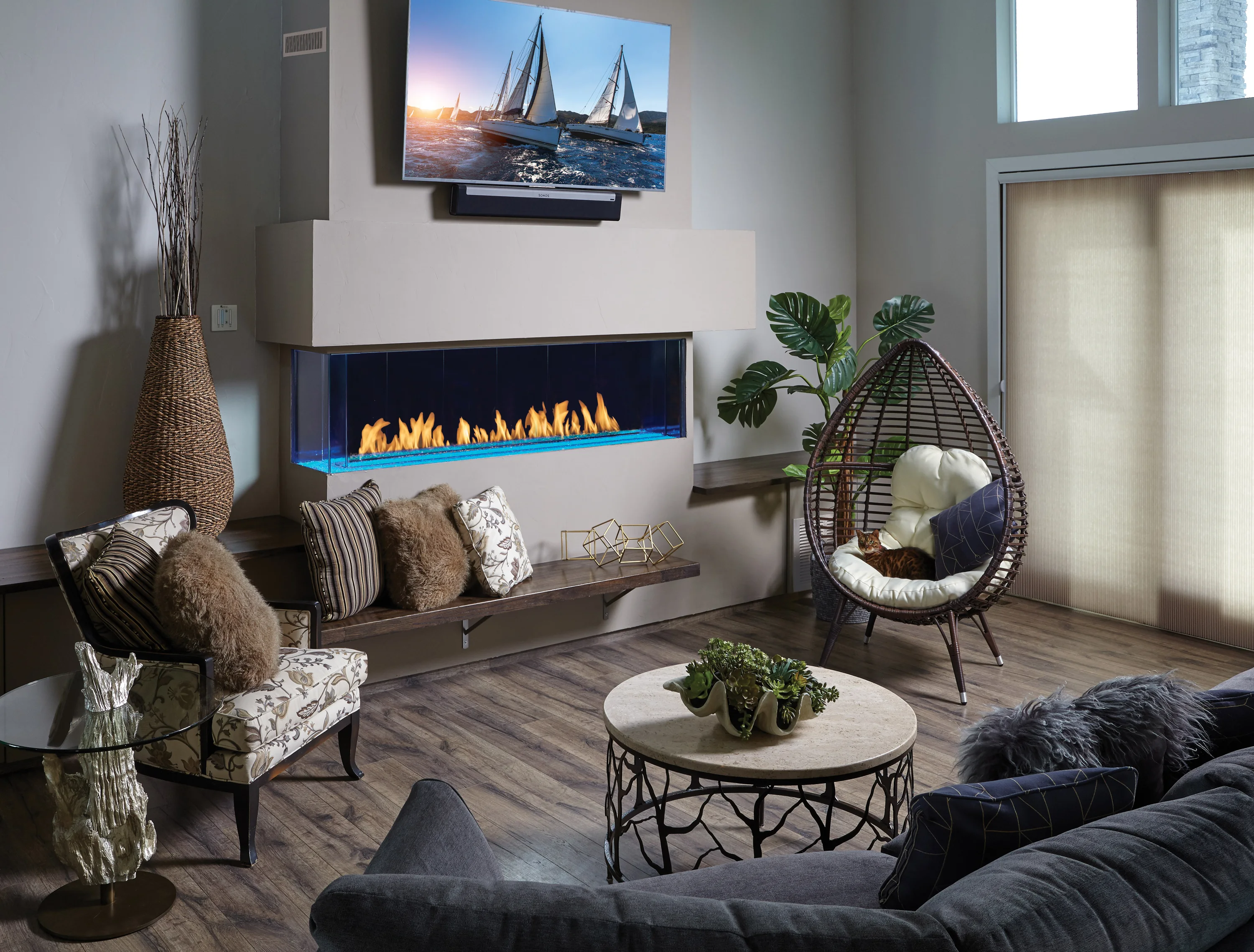 Modern living room with a wall-mounted TV above a contemporary fireplace. Room features a hanging chair, cushioned benches, potted plants, glass-top table, and a round coffee table with plants.