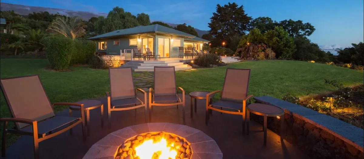 Patio with five chairs and a table surrounding a lit fire pit, set in a garden with a house in the background at dusk. The house has lit windows and a porch with chairs.
