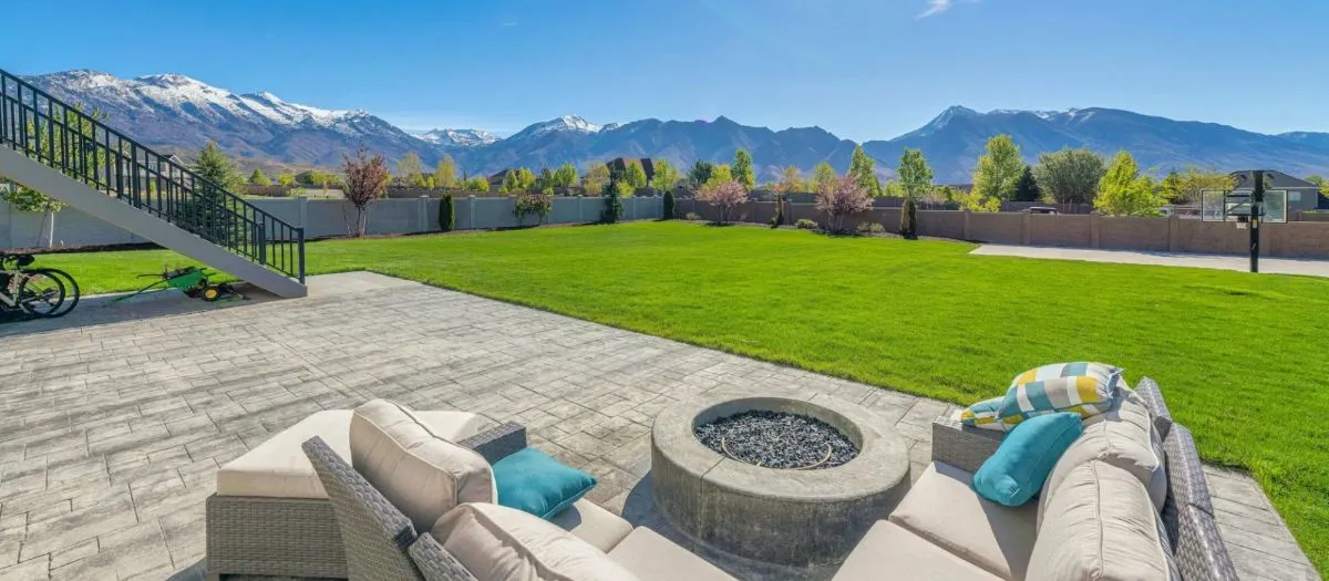 Patio with outdoor seating and a fire pit overlooking a spacious green lawn, with a backdrop of snow-capped mountains under a clear blue sky.