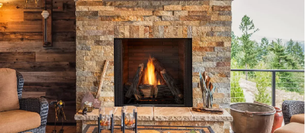 Outdoor stone fireplace with a fire burning inside. The patio features a tiled floor, wicker armchairs, and scenic views of trees and distant mountains in the background.