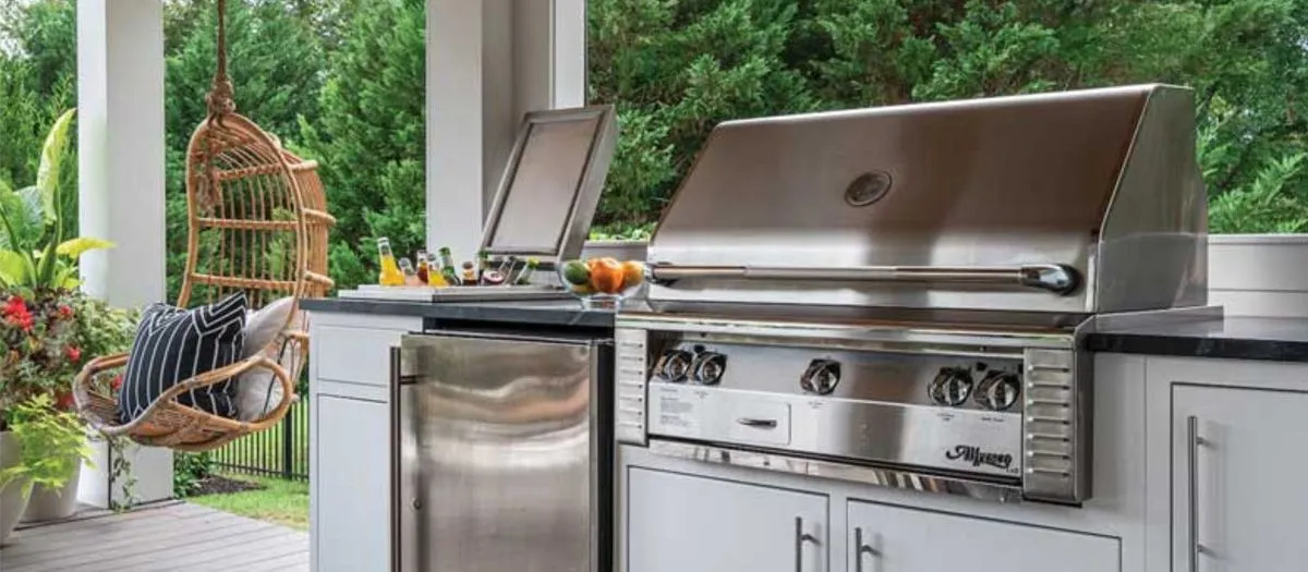 Outdoor kitchen with stainless steel grill, mini fridge, counter space with drinks, and a hanging wicker chair with striped cushions. The area is surrounded by greenery and plants.
