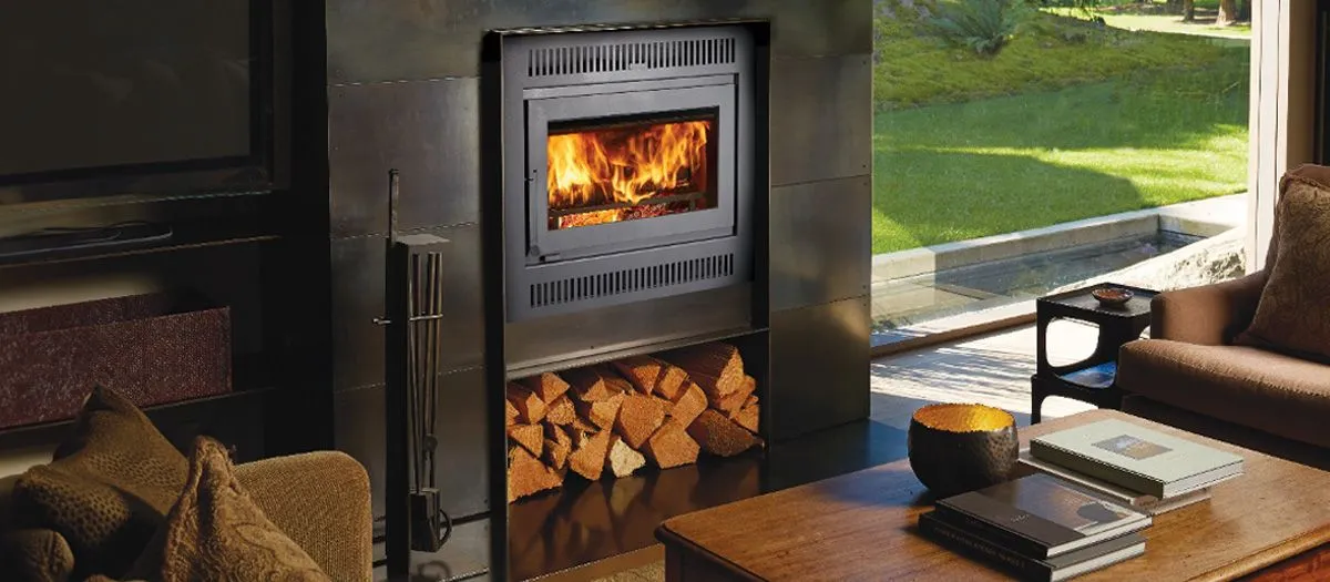 A cozy living room with a modern fireplace burning wood, firewood stocked underneath. Nearby are a brown couch, a wooden coffee table with books, and large windows revealing a green lawn outside.