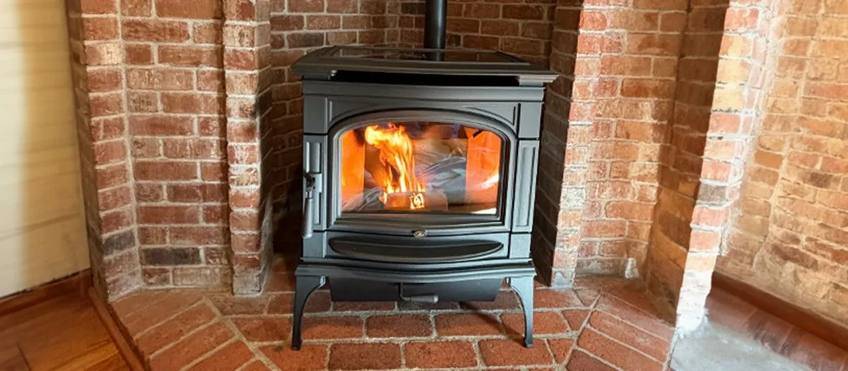A black wood-burning stove with a visible fire inside, set within an arched brick alcove. The floor is made of red, hexagonal tiles.