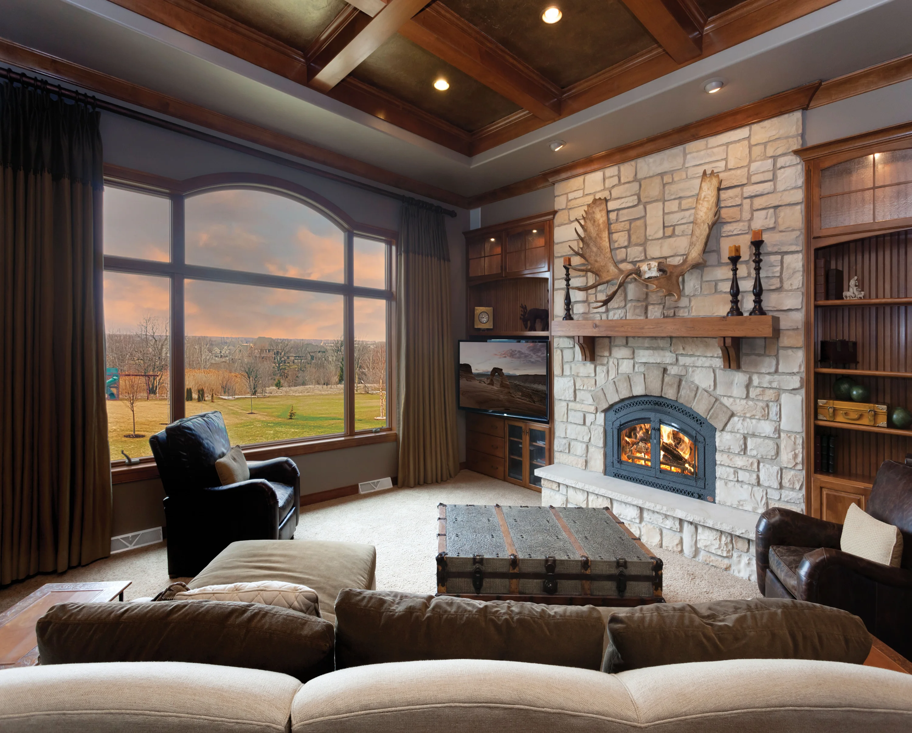 Cozy living room with stone fireplace and mounted moose antlers, a lit fire, large arched window overlooking yard, leather chairs and trunk coffee table