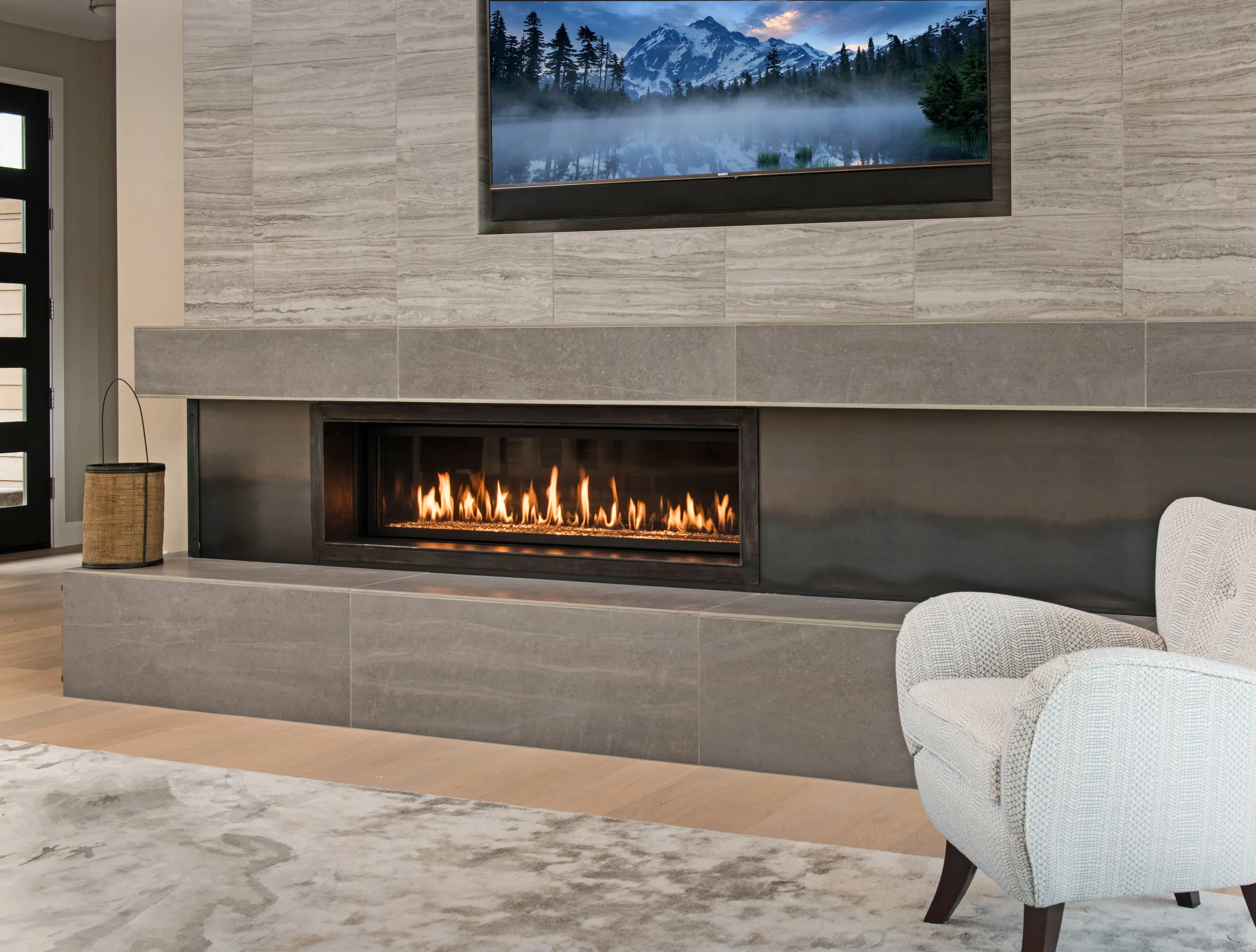 Modern living room with long rectangular gas fireplace set into gray stone tile wall, TV above, woven basket and light upholstered chair nearby